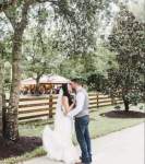a bride and groom standing on a walk with trees, a fence and a pavilion in the background